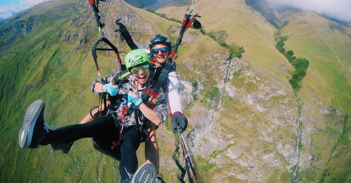 Two people tandem paragliding over green hills and valleys near Wānaka, with excited expressions and a selfie stick.