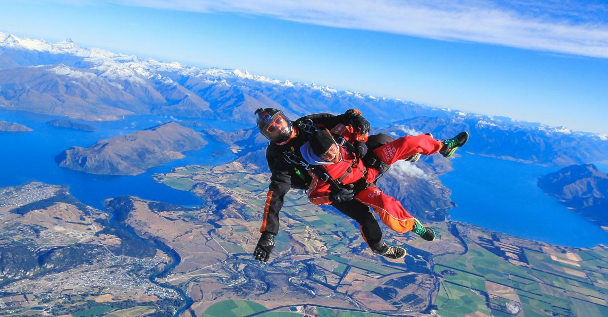 Tandem skydivers freefalling above Lake Wanaka with snowy mountains and patchwork farmland below.