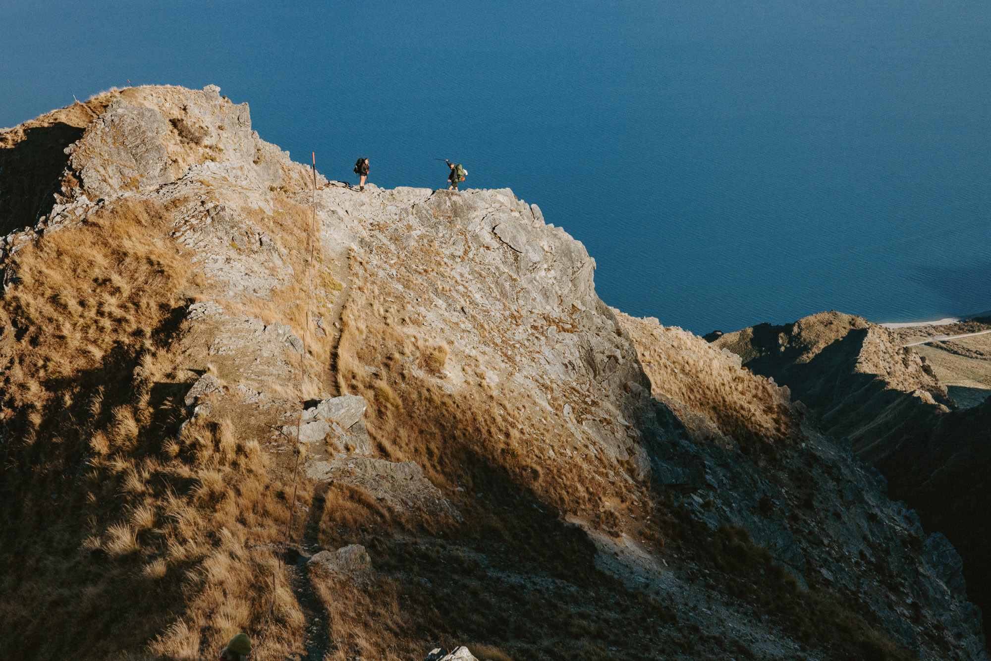Hikers navigating a rocky ridgeline above Lake Wānaka with sweeping views and golden alpine terrain.