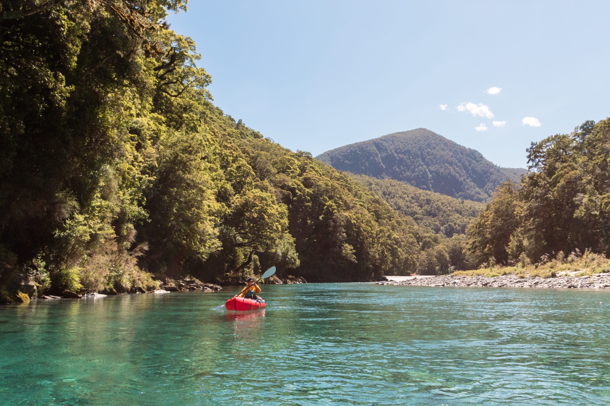 Person paddling a red packraft down a clear turquoise river surrounded by lush native forest in Makarora, New Zealand.
