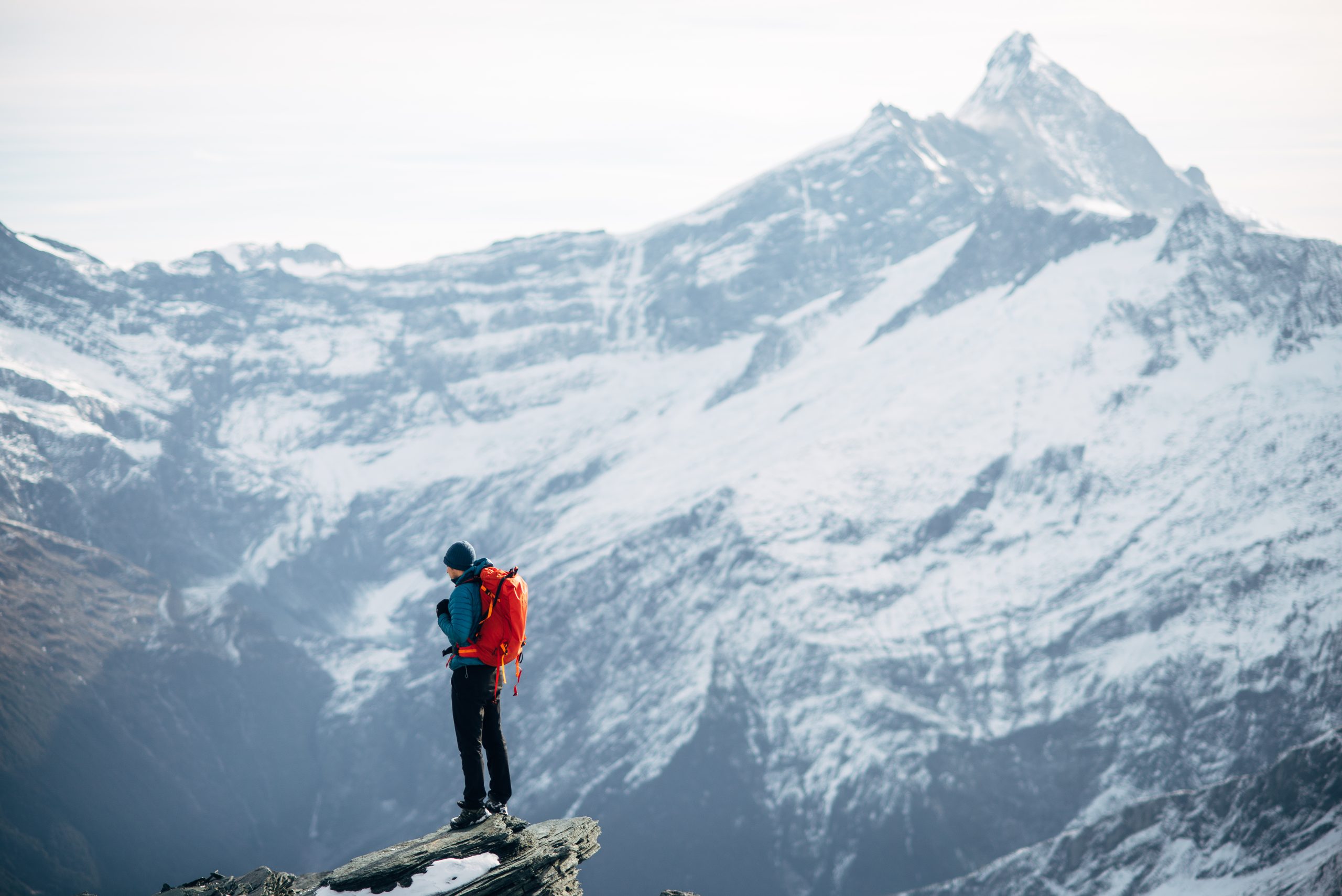 Solo hiker with a red backpack standing on a rocky ledge, looking out over snowy peaks in Mount Aspiring National Park.