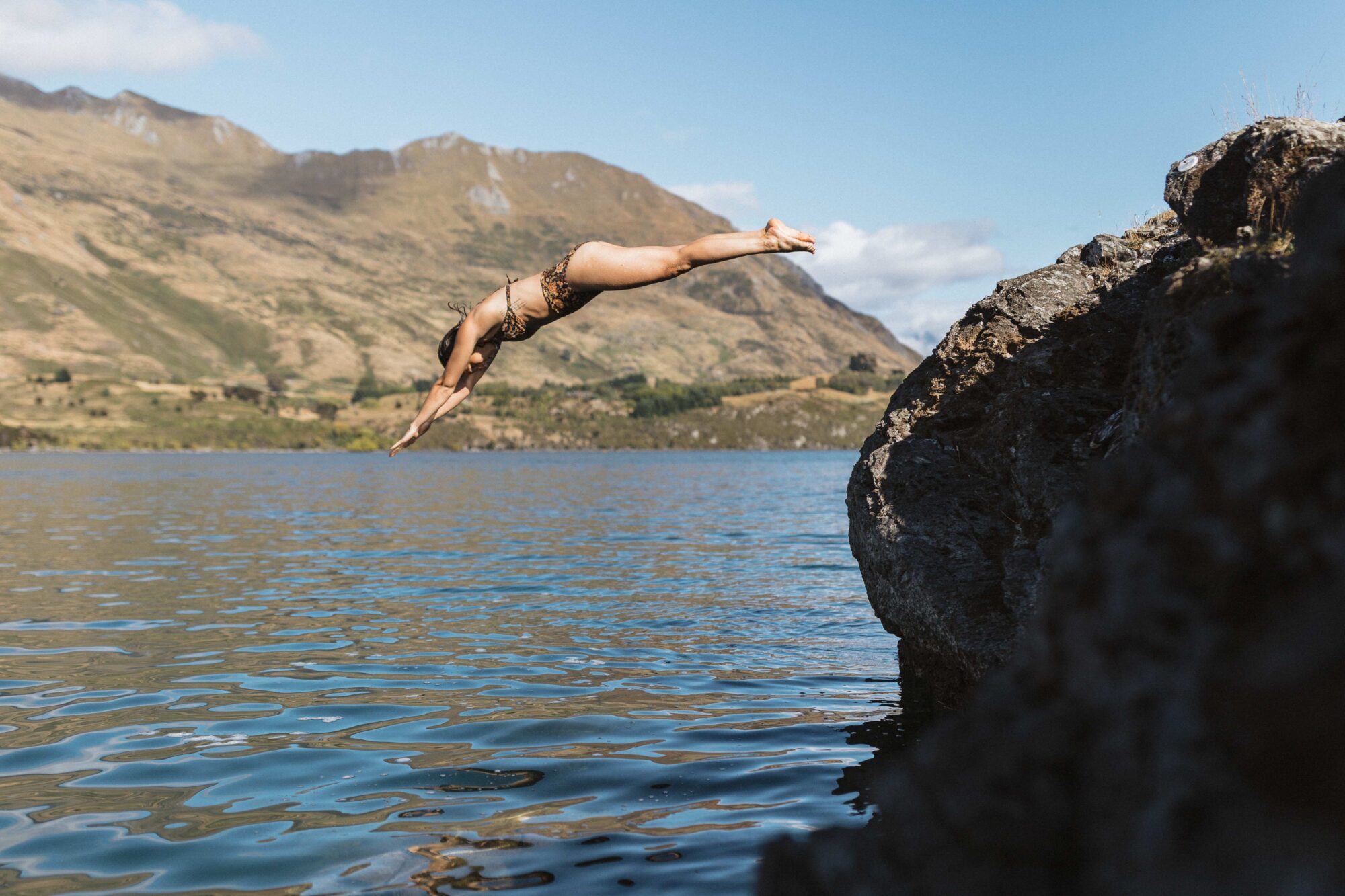 Swimmer leaping off rocks into the clear water at Ruby Island, Lake Wānaka